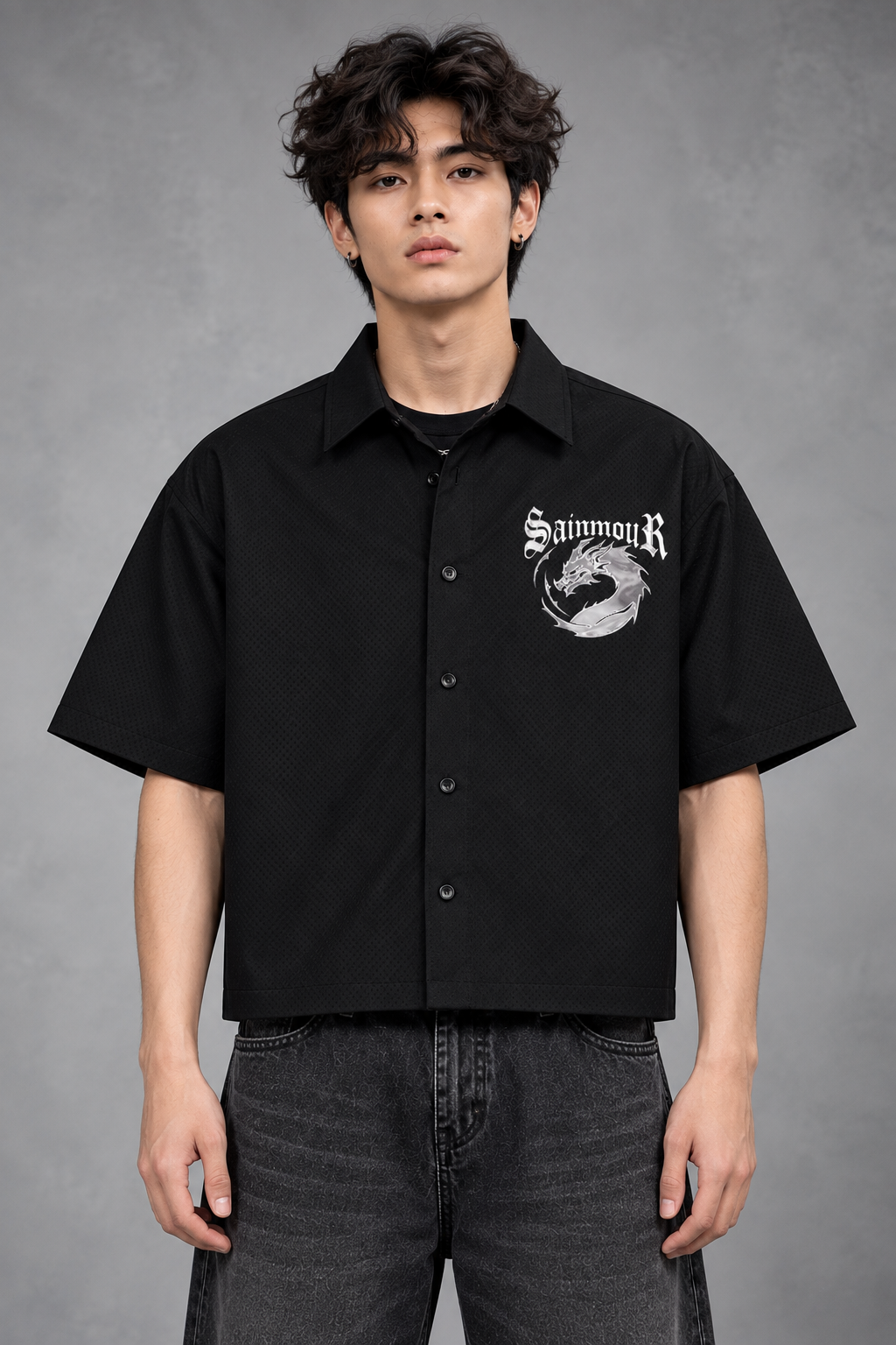 A man wearing a black short-sleeve button-up shirt with a white 'SainmiR' graphic on the left chest, paired with dark gray jeans, standing against a textured gray studio background.