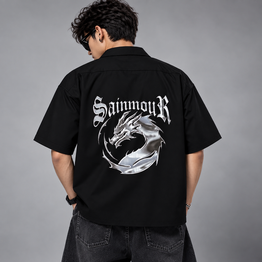 A man viewed from behind wearing a black short-sleeve button-up shirt with a large metallic silver dragon graphic and the word 'SaimouR' across the back, paired with dark jeans against a gray studio background.