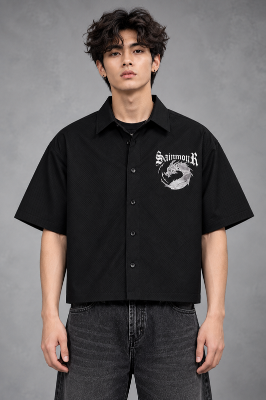 A man wearing a black short-sleeve button-up shirt with a white 'SainmiR' graphic on the left chest, paired with dark gray jeans, standing against a textured gray studio background.