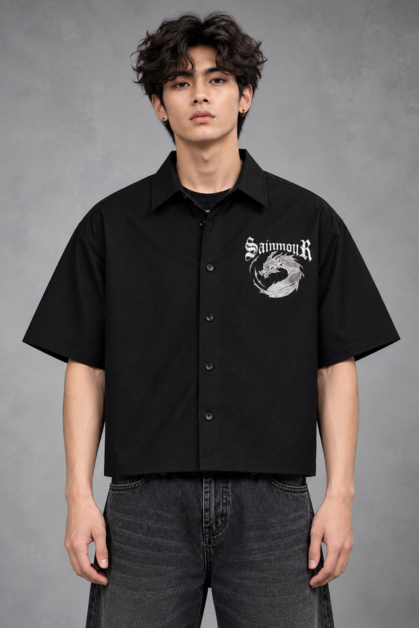 A man wearing a black short-sleeve button-up shirt with a white 'SainmiR' graphic on the left chest, paired with dark gray jeans, standing against a textured gray studio background.